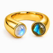 A polished gold open cuff ring featuring two oval cabochon stones, one with a blue-white iridescent sheen and the other with a blue-green sheen, set against a plain white background.