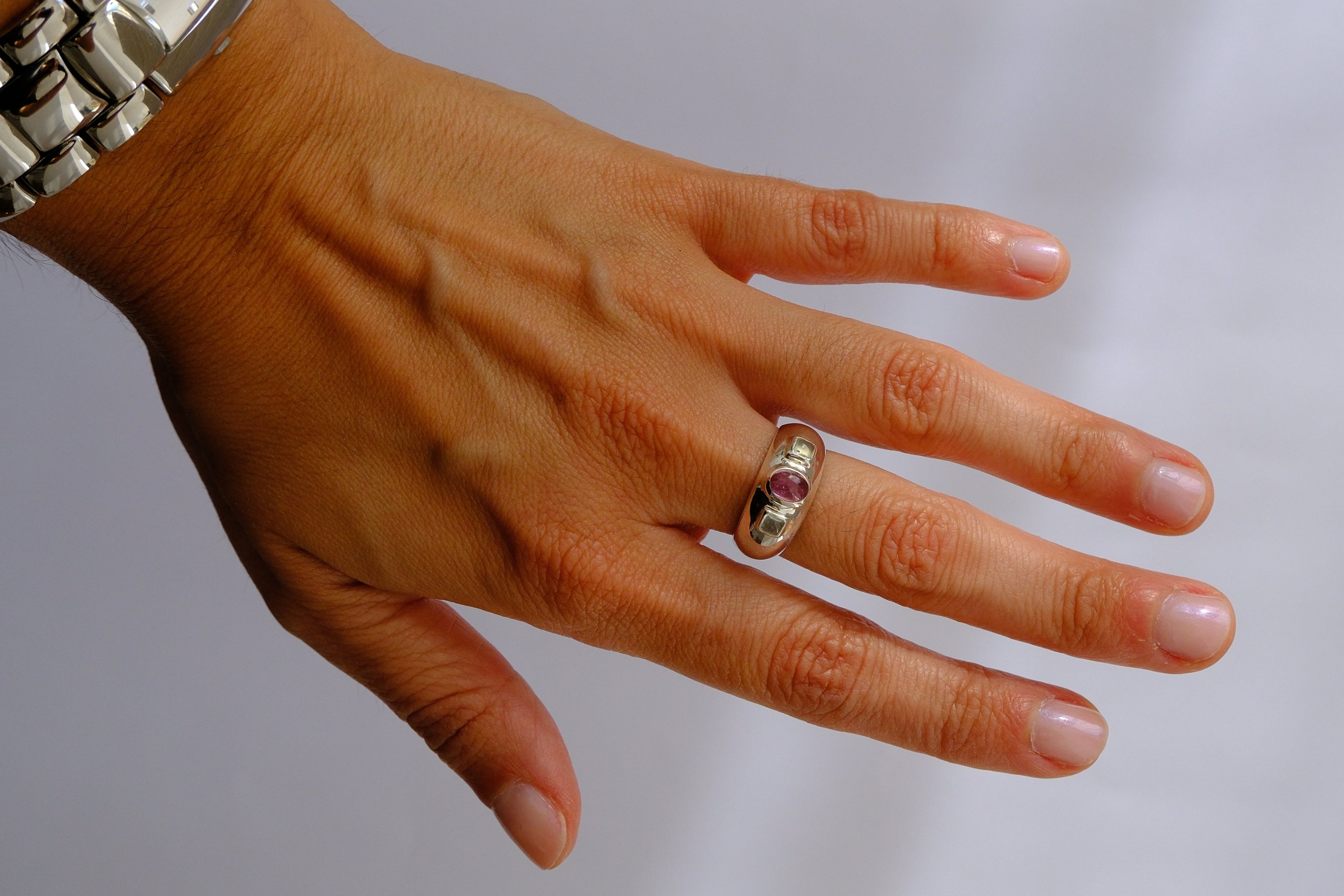 A silver band ring with small clear stones worn on a person's ring finger, shown against a plain light gray background. The person's hand and wrist, along with a silver watch, are visible.