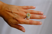 A silver-toned ring with a large oval light green gemstone and a halo of small clear stones, shown worn on a person's ring finger. The person's hand and a matching silver link bracelet are also visible against a soft light gray background.