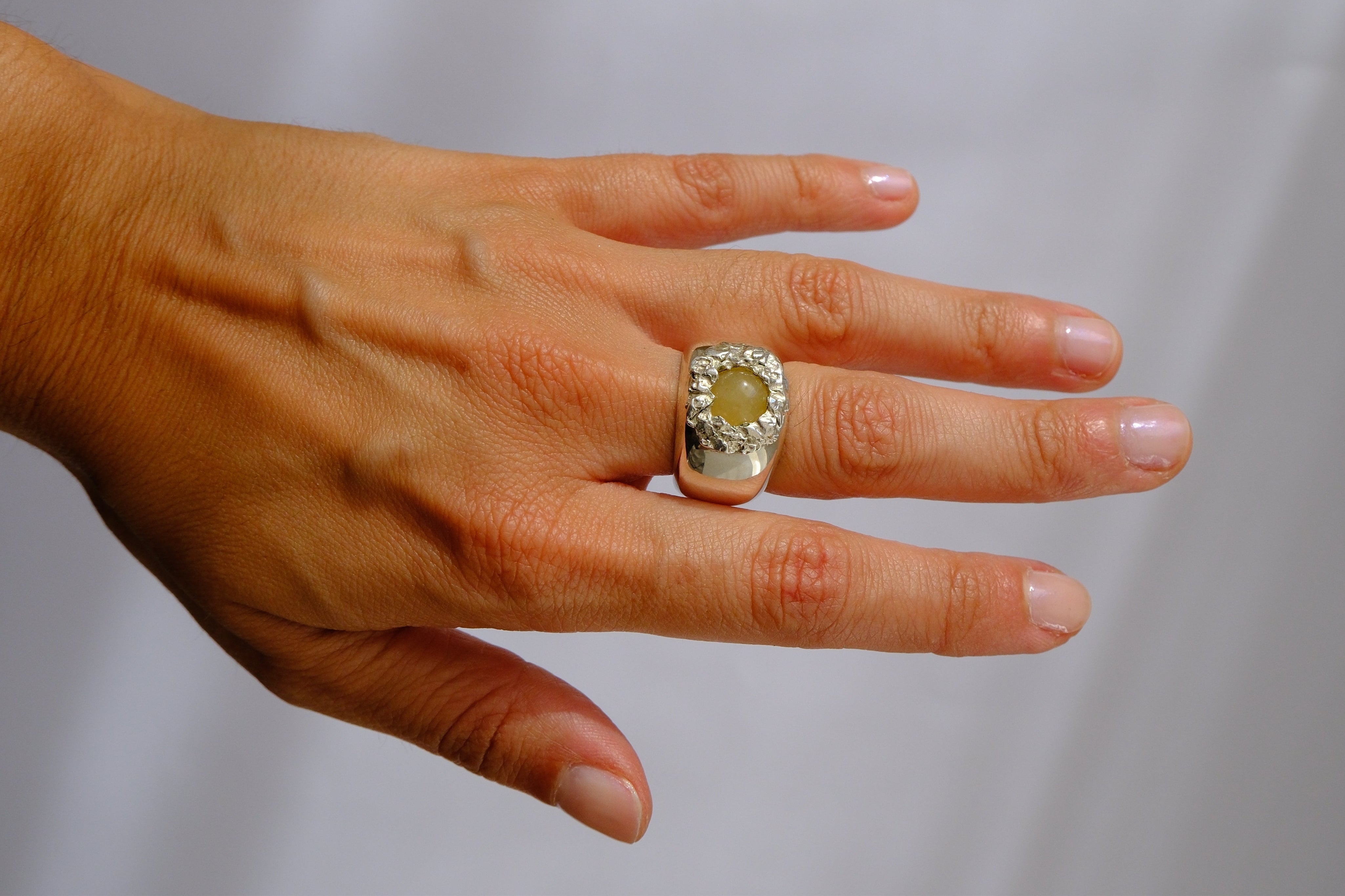 A silver-toned ring with a large oval light green gemstone and a halo of small clear stones, shown worn on a person's ring finger. The person's hand and a matching silver link bracelet are also visible against a soft light gray background.