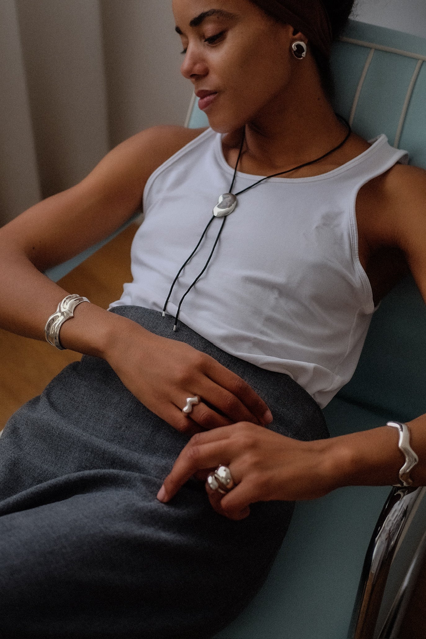 Woman sitting on a couch wearing a white tank top and gray pants, with jewelry on.