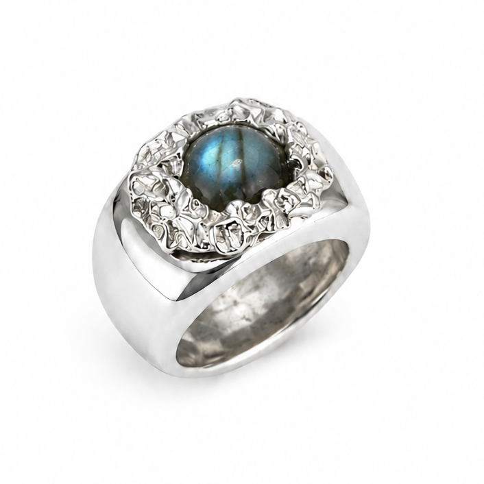 A silver-toned ring featuring a round iridescent blue-green center stone surrounded by a halo of small clear stones, set on a plain white background.